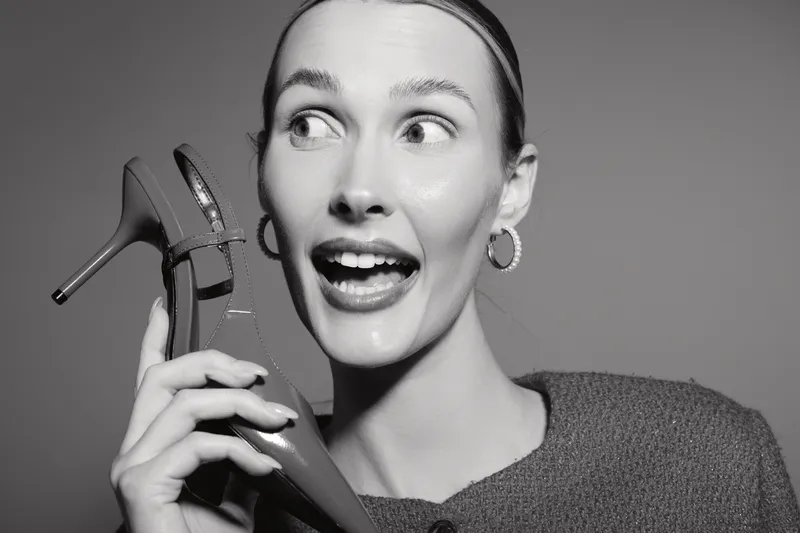 Young white woman with light skin holds red high heel shoe near face and looks surprised. Close-up portrait in black and white studio. Concept of fashion and emotions.