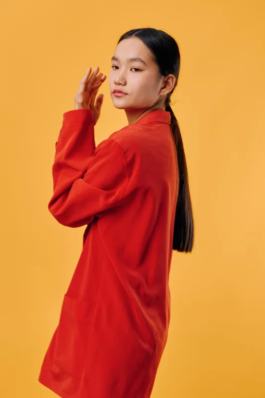 Young woman with long hair wearing a red jacket posing with serious and confident expression on orange background in studio. Female model standing sideways with hand near face.