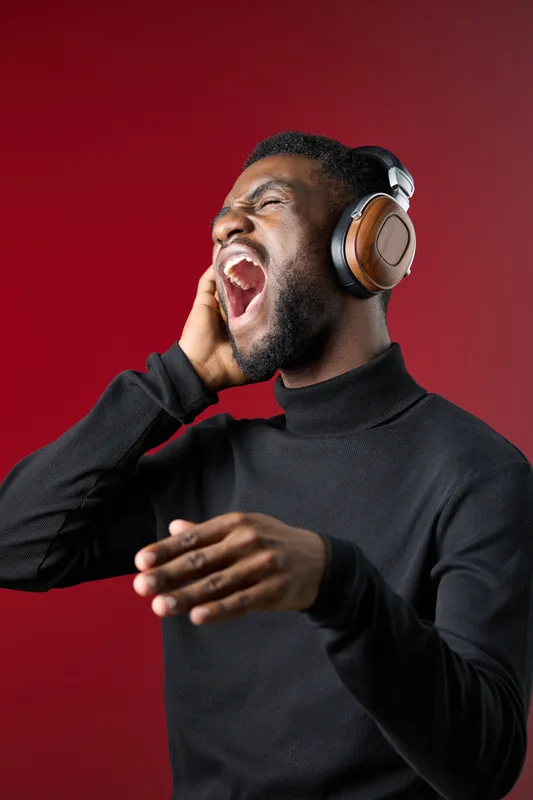 A joyful Black man wearing stylish headphones passionately singing with eyes closed against a vibrant red background, expressing happiness and music s power