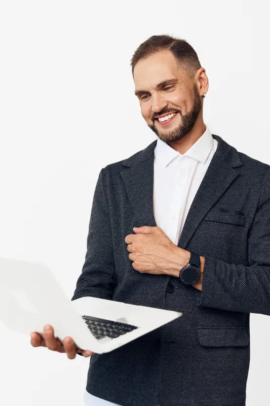 A professional man stands against a solid colored background, projecting confidence and focus as he gestures with a smile while presenting with a laptop in hand.
