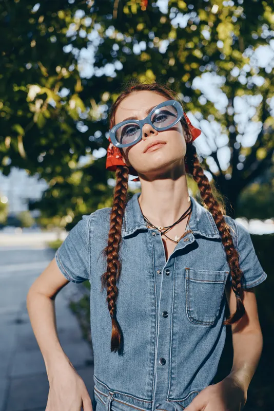 Lifestyle woman in modern boho-western denim street style with braided hair and oversized blue sunglasses, captured in warm film color tones under natural sunlight.