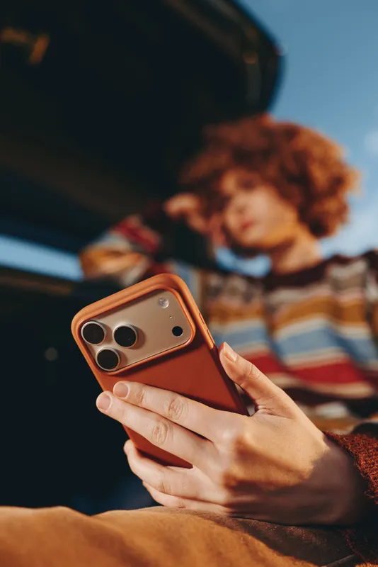 Woman holding smartphone, hands visible, curly hair person blurred in background, indoor home lifestyle on sofa, mobile technology for communication, relaxed texting mood, soft light