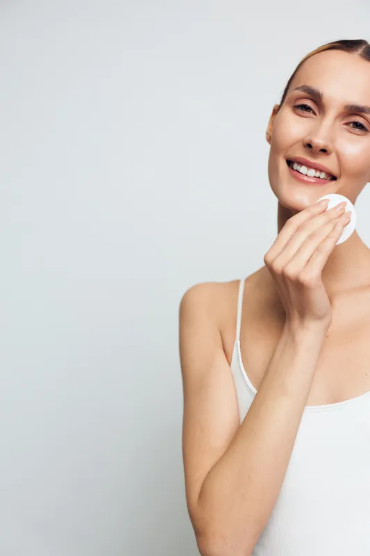 Smiling woman with clear skin using cotton pad to remove makeup on white background. Concept of skincare, beauty routine, cleansing, health, and self-care with natural light.