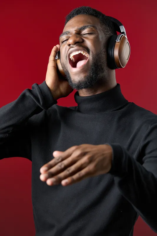 A young African American man passionately singing while wearing headphones against a vibrant red background, showcasing a joyful emotion and modern fashion