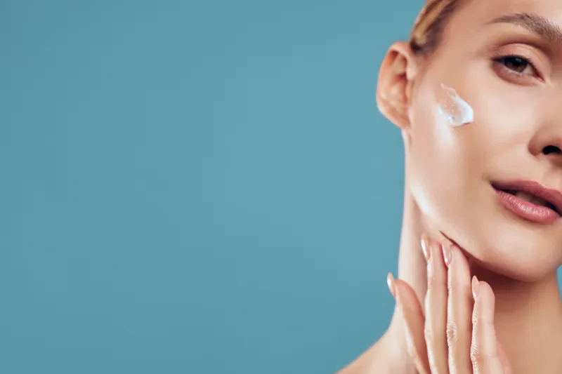 Young woman applies cream to her cheek with a calm focused expression, isolated against a blue studio backdrop, showcasing skincare routine and healthy glowing skin