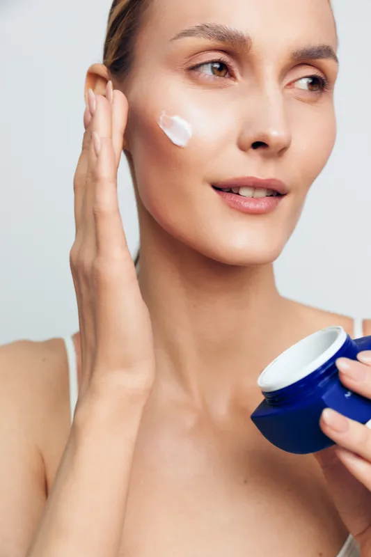 Woman with clear skin applying white face cream on cheek, holding blue jar. Close-up on white background. Concept of skincare, beauty routine, moisturizing, and self-care with natural light.