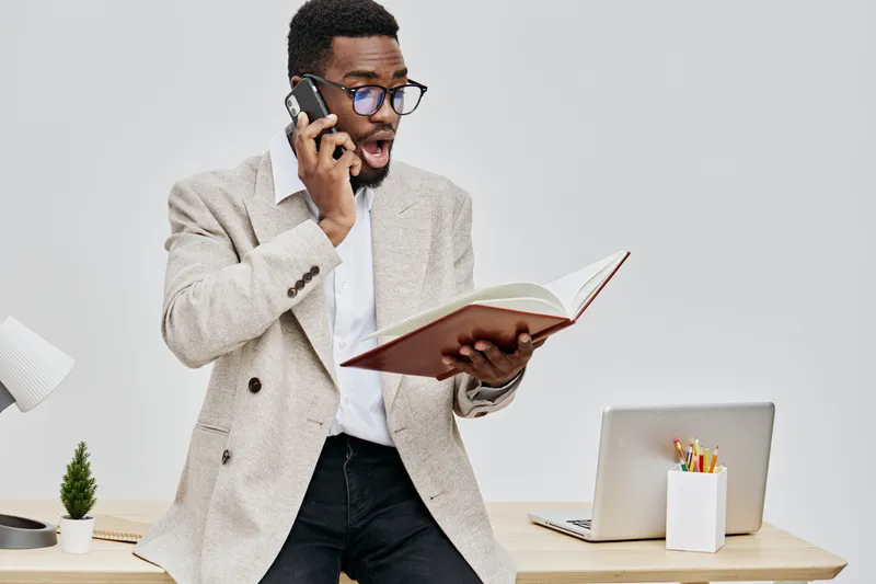Businessman talking on smartphone while reading a book at a modern office desk, with laptop and stationery, portraying professional communication and multitasking in a corporate environment. People li