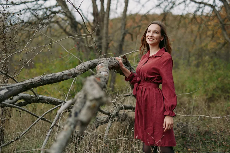 An authentic portrait of a woman in a crimson dress beside a fallen branch, set in a quiet natural scene that radiates warmth, sincerity, and subtle autumn charm.
