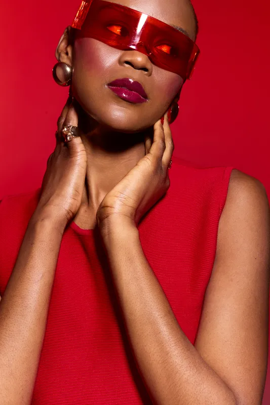 Stylish black woman with dark skin wearing red sleeveless top and red sunglasses poses against red background. Concept for fashion, beauty, and confident female model.