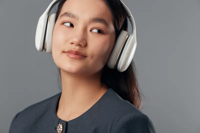 Young woman listening with headphones, looking sideways with thoughtful expression in studio. Female person with long hair and casual outfit on gray background with soft lighting.