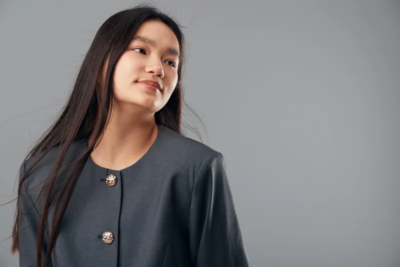 Young woman with long dark hair wearing a grey buttoned jacket standing and looking to the side against a plain grey background in studio lighting.