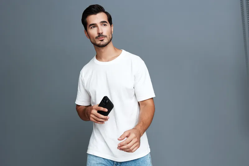 Casual young man standing against a gray background, holding a smartphone while looking thoughtfully to the side. Modern lifestyle concept.