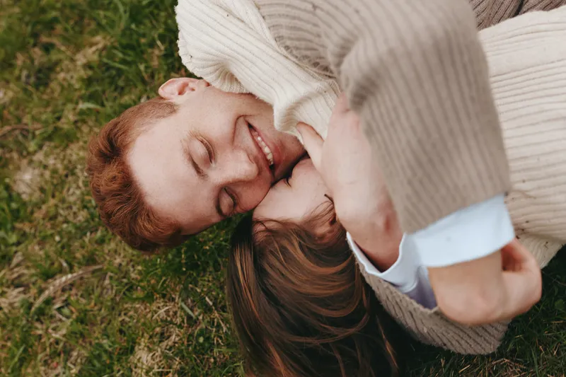 A couple lies on the grass, smiling and embracing under a knit blanket, capturing intimate outdoor affection. A lifestyle portrait of romance, connection, and happiness in a sunny, natural setting.
