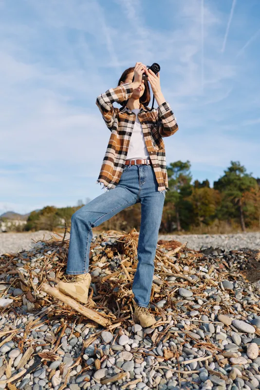 Photographer in casual outdoor gear on rocky shore, plaid shirt, denim jeans and boots, capturing a landscape moment with camera against a clear sky and autumn trees.