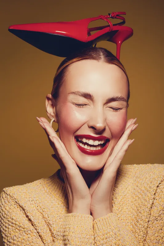 Happy woman with closed eyes and red lipstick smiles joyfully while balancing a red high heel shoe on her head. Concept of fun, fashion, and playful mood in studio.