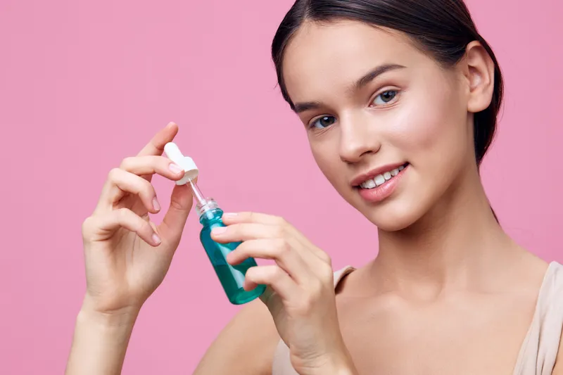 Young woman applying skincare product with blue serum on pink background, showcasing healthy skincare routine and beauty concept