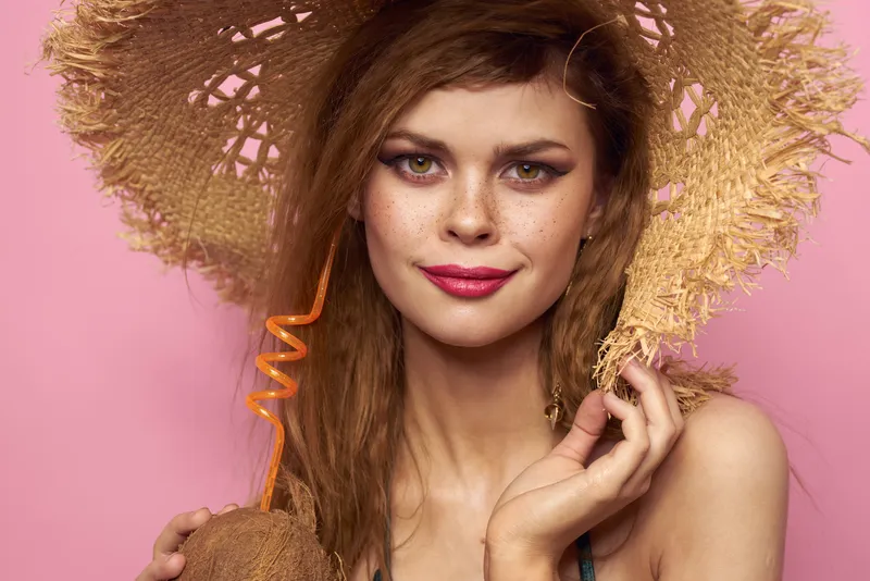 Женщина держит в руках коктейль в кокосе, на ней купальник и пляжная шляпа, рядом косметика