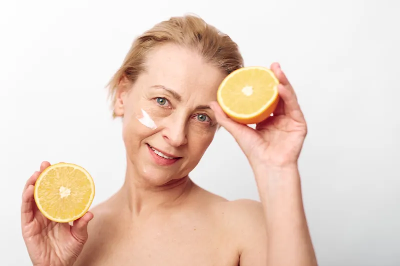 Middle aged woman with clear skin holds two halves of orange fruit near her face with cream on cheeks. Bright skincare concept, healthy lifestyle and natural beauty on white background.
