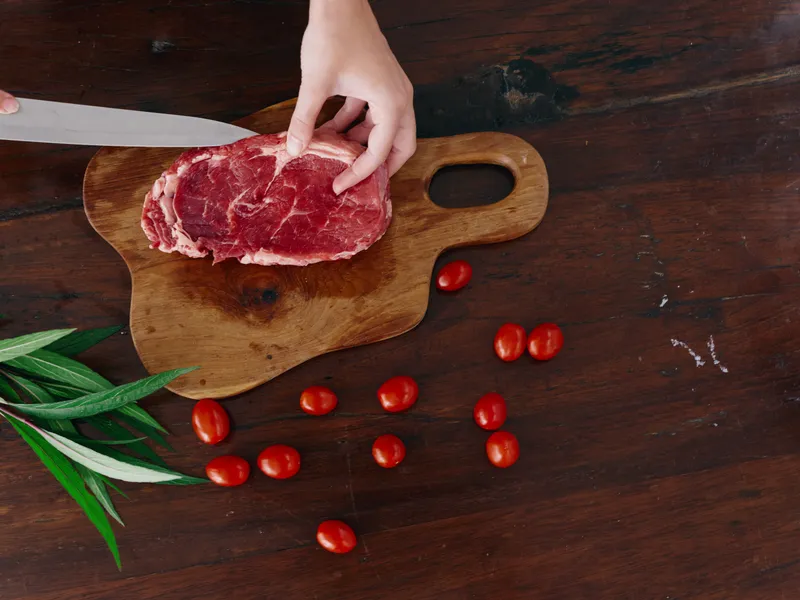 Женщина с ножом в руке нарезает свежее стейковое мясо для жарки на кухне, рядом на столе соль, перец и другие специи, красные помидоры черри и зелень