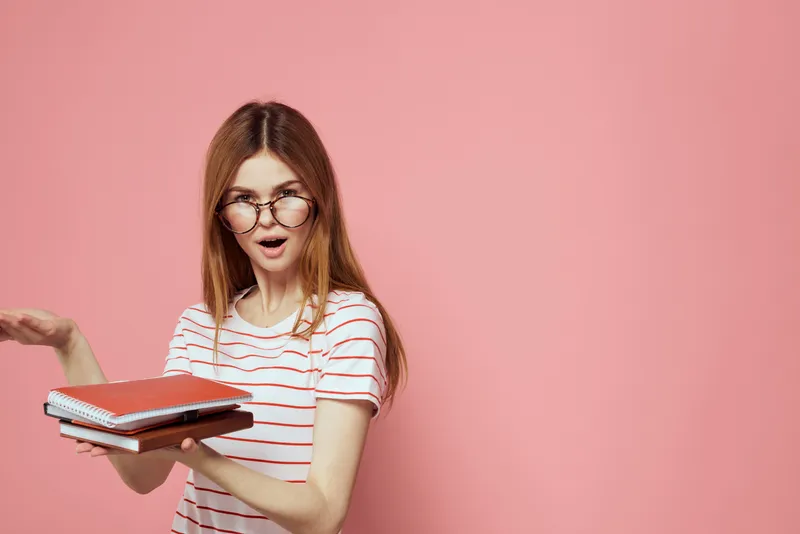 Молодая студентка с книгами на розовом фоне, в очках, крупный план лица