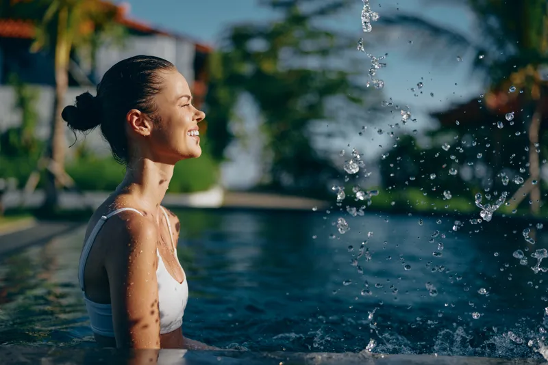 A joyful woman relaxes in a pool, water splashes around her as bright sunlight catches her smiling face, a serene lifestyle moment highlighting outdoor leisure, wellness, summer escape, and carefree