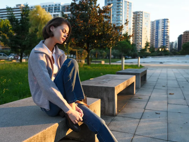 A reflective woman sits on a park bench beside a quiet urban setting. She adjusts her shoe, lost in thought, conveying calm, focus, and casual style during golden hour.