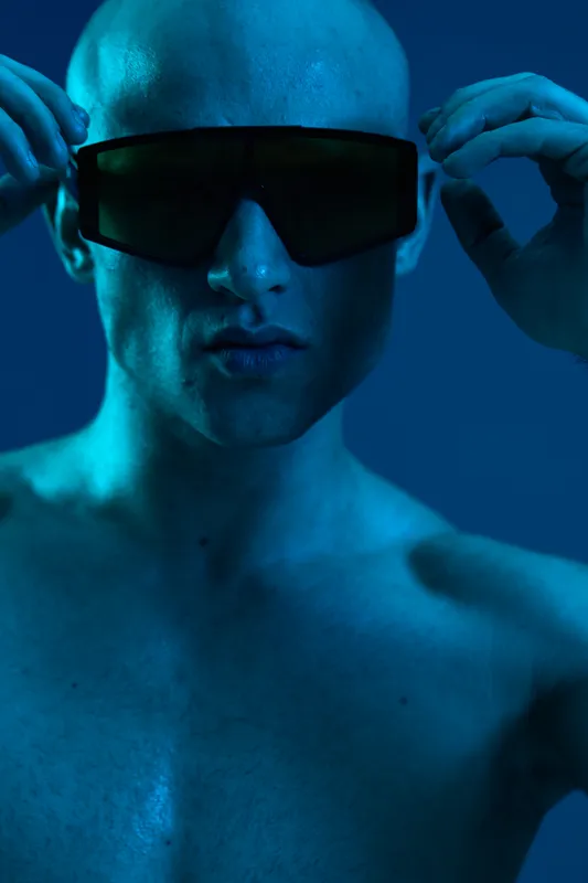 Young man adjusting sunglasses, showcasing a striking blue light in a moody atmosphere. This captivating image reflects modern fashion and style.
