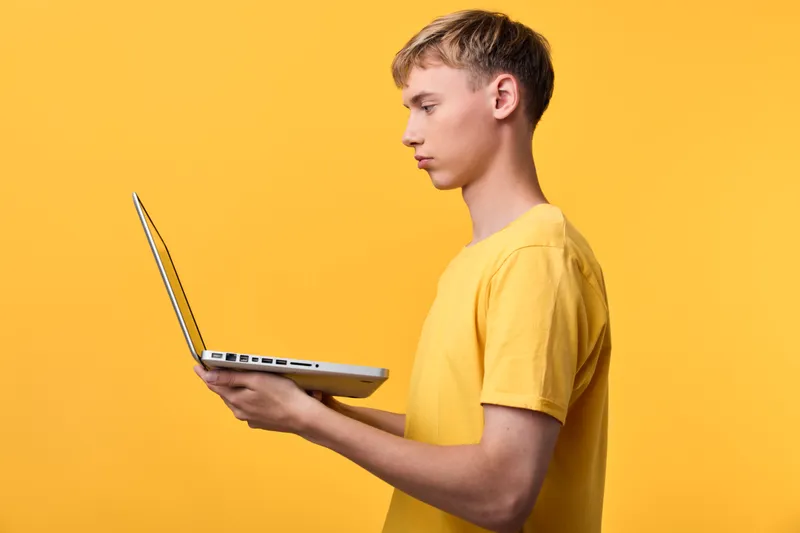 Young man using a laptop against a bright yellow background, showcasing technology, concentration and learning through digital devices in a casual studio setting