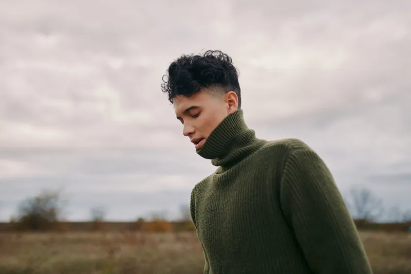 An honest, candid portrait of a thoughtful man in a thick green turtleneck, underscoring authenticity through quiet emotion against a soft outdoor sky.