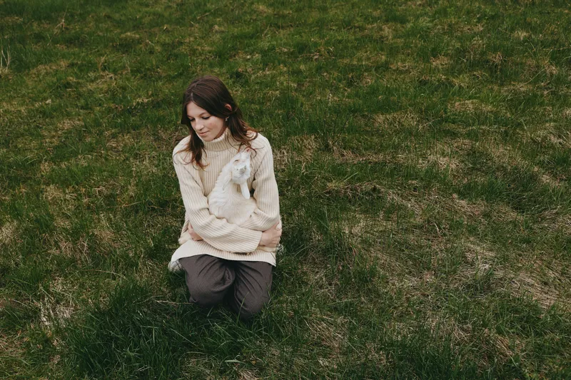 A woman kneels on lush grass, cradling a small goat kid in her arms in a rural setting. Bright natural light highlights a serene moment of connection, care, and everyday animal companionship.