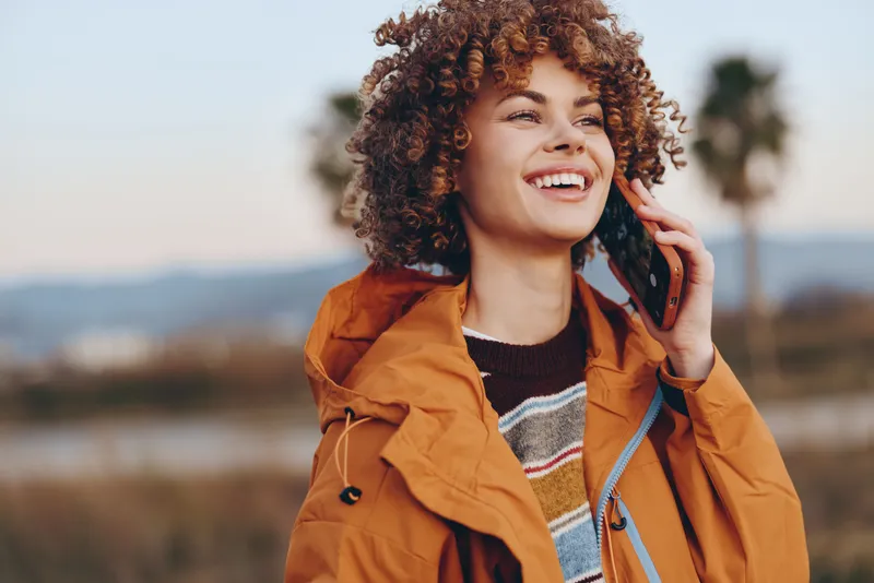 Woman lifestyle portrait outdoors, curly hair, smiling young woman in orange jacket, hand by ear talking on phone, sunset landscape, soft bokeh, casual autumn travel, warm light, weekend