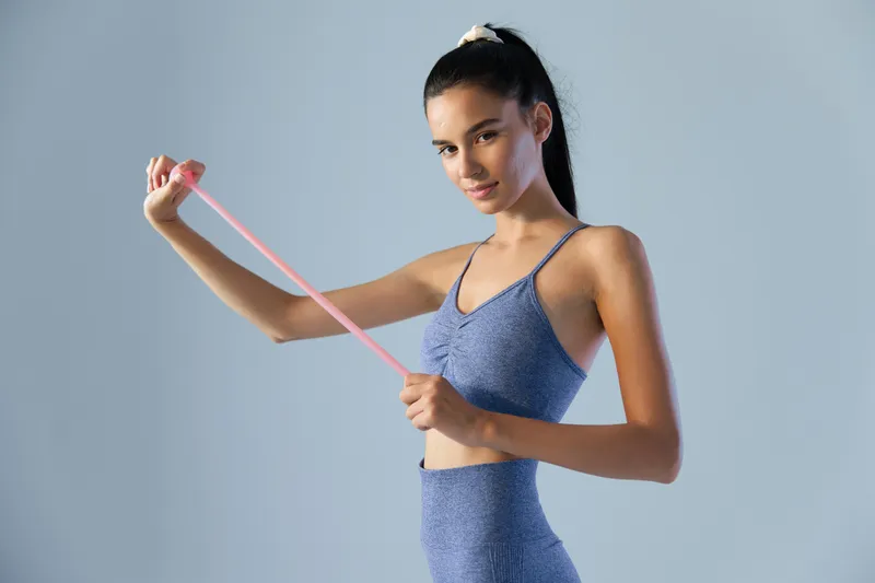 young woman sport fitness stretching resistance band in blue activewear against plain background, demonstrating strength and focus during workout session