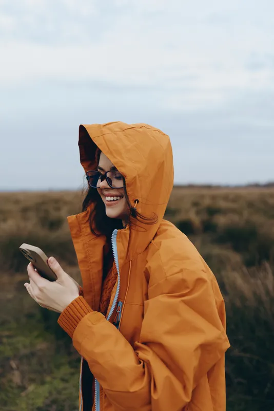 Smiling woman lifestyle outdoors in rural nature, wearing orange hooded jacket, holding smartphone and scrolling, cloudy sky and field landscape, casual travel mood for everyday photo