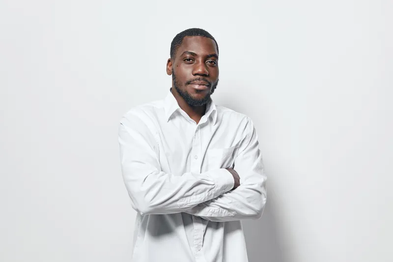 African American man with dark skin wearing white shirt standing with arms crossed against white background. Confident male portrait with casual style and serious expression.