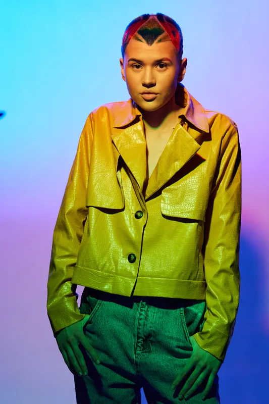 young woman with colorful hairstyle wearing a shiny yellow jacket and textured pants stands confidently against vibrant gradient background under dramatic lighting