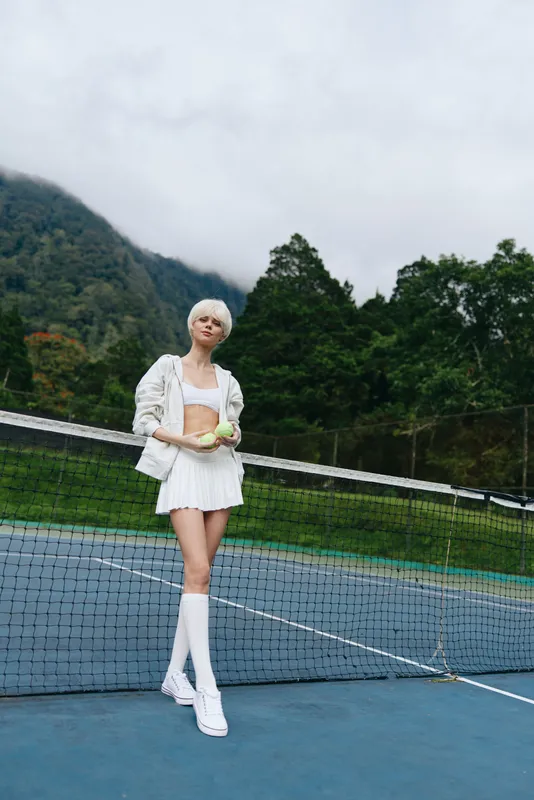 Young woman in sporty outfit holding a tennis ball on a court, exuding confidence and energy in a lush green outdoor setting