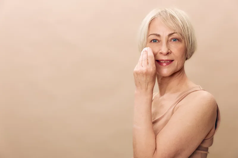 Mature woman with short blonde hair gently cleansing her face with a cotton pad on beige background. Concept for skincare, beauty routine, anti aging and self care.