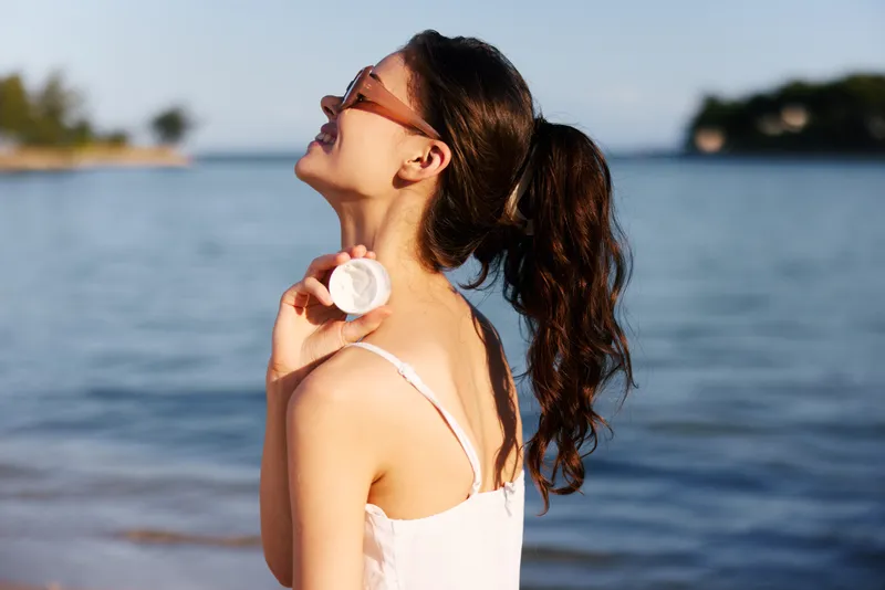 Smiling woman applying sunscreen by the beach, enjoying sunny summer day with sunglasses and casual white dress, natural skincare and outdoor relaxation.