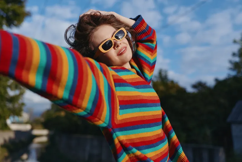 Woman smiling in a colorful striped sweater and yellow sunglasses outdoors, curly hair blowing, casual outfit, joyful pose with outstretched arms on a sunny travel lifestyle moment.