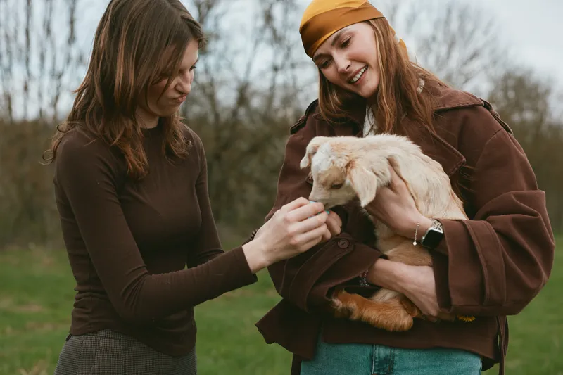 Two women outdoors petting a fluffy goat kid in a casual countryside setting, smiling and relaxed, capturing a warm lifestyle moment of connection with animals showcasing warmth and joy.
