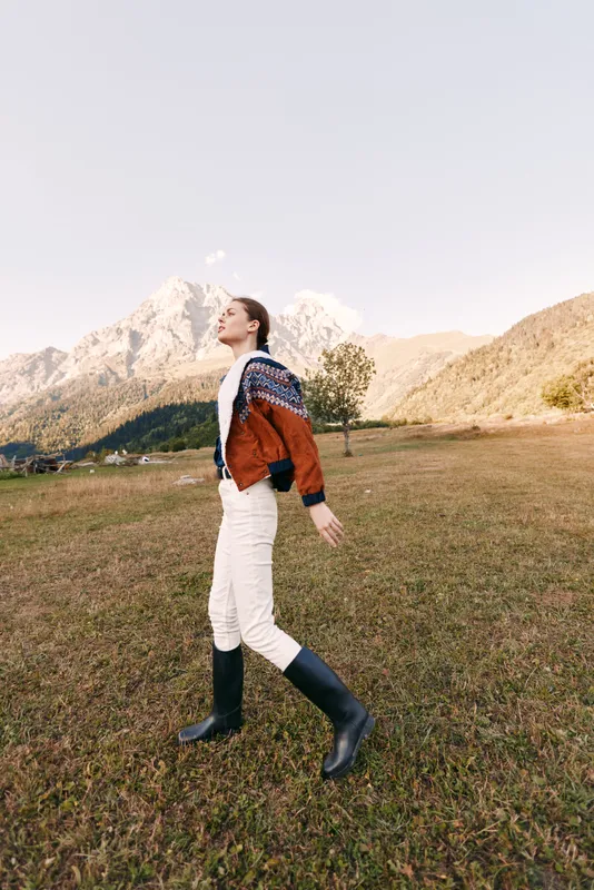 Woman walking in mountains field wearing a cozy sweater and boots outdoors in autumn, casual jacket and knitwear style captured in a serene landscape meadow travel portrait.