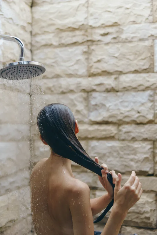 Woman washing long hair under outdoor shower with natural stone wall background in warm daylight, relaxing skincare moment with water drops.
