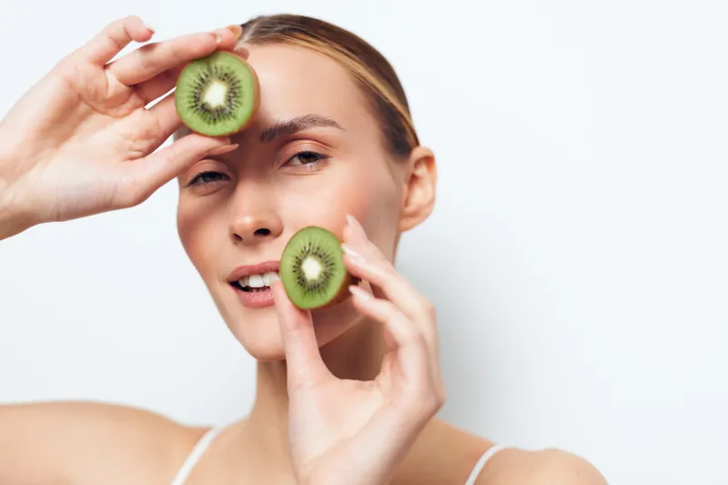 Young woman holds two halves of kiwi fruit near face with gentle smile, natural skin, isolated on white background, healthy lifestyle concept, fresh fruit closeup.