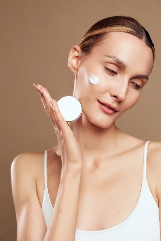 Woman applies white face cream on cheek, closeup portrait for skincare beauty selfcare wellness concept, minimal studio lighting, warm brown background, soft glow