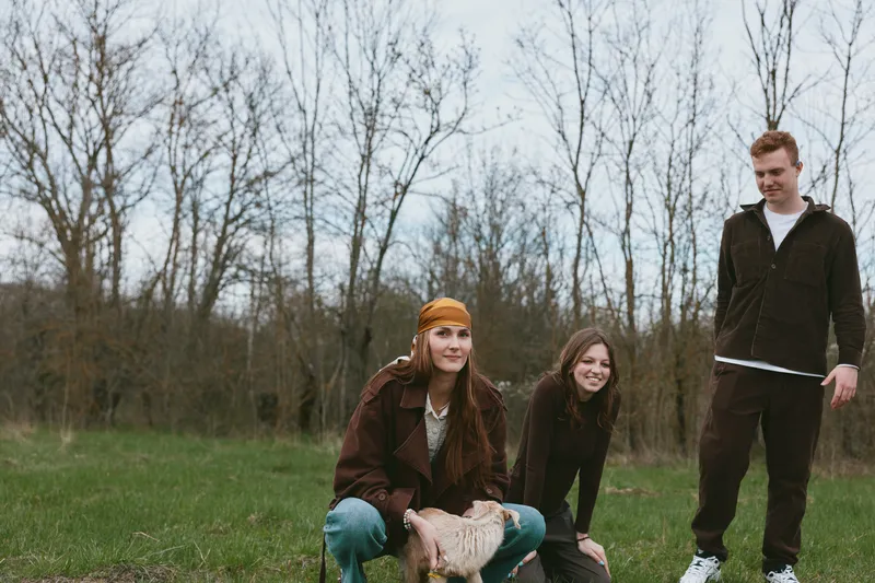 Three young adults share a playful moment with a goat kid in a grassy park, casual clothing, natural light, candid lifestyle shot capturing friendship, rural charm, and simple outdoor joy.