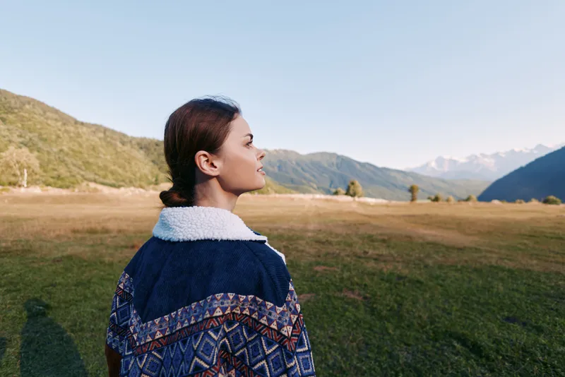 Woman in patterned jacket stands outdoors in a meadow, profile view facing mountains and expansive landscape. Nature travel scene with serene sky and calm countryside mood.
