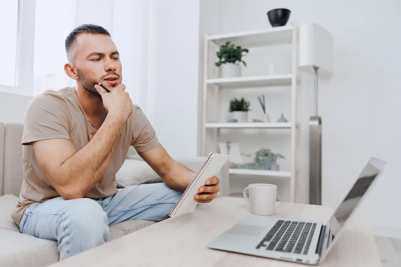 Thoughtful man contemplating ideas at home while sitting on a couch with a notebook and laptop, creating an inspiring atmosphere for productivity and creativity.