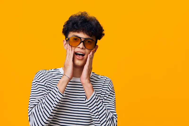 surprised young man wearing striped shirt and orange sunglasses expressing shock with hands on face against vibrant yellow background, casual style