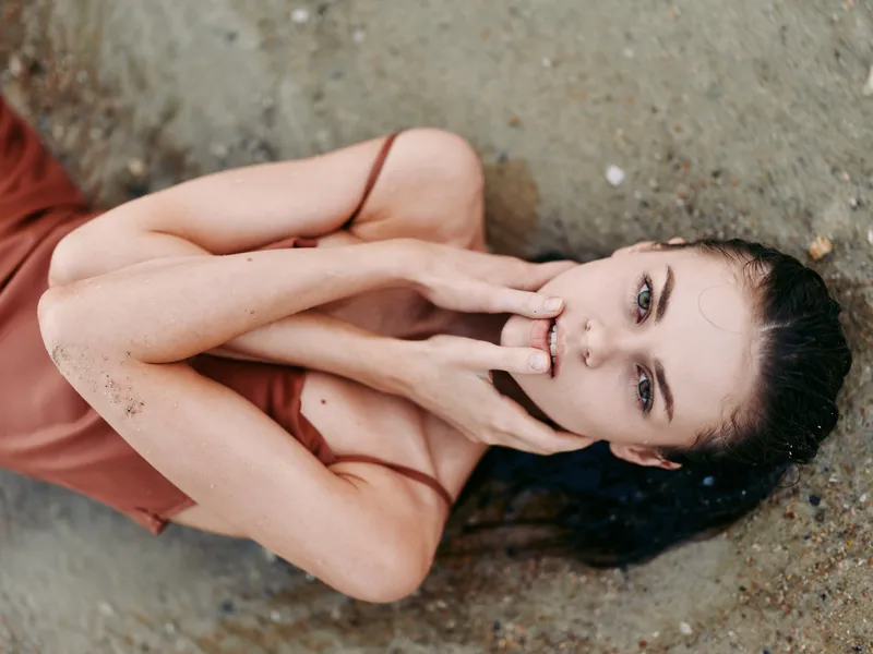 A woman lies on sandy ground in a rust dress, hands softly near her lips, eyes gazing with calm intensity, wind tousled hair, an intimate beach portrait.
