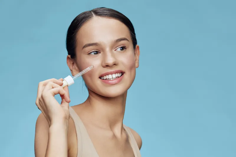 Young woman applying skincare serum with a dropper, smiling against a soft blue background, emphasizing healthy skin and beauty